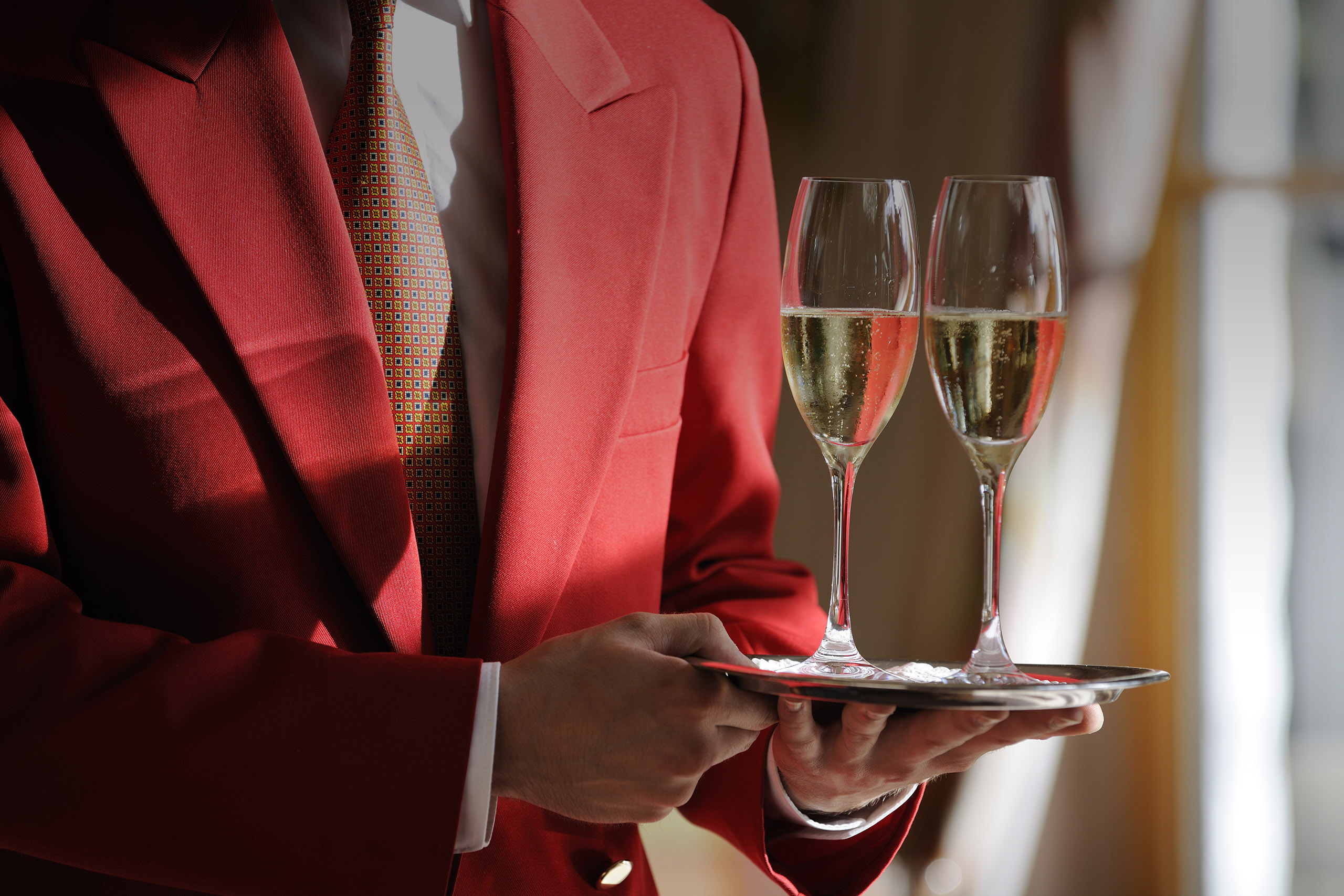 Ein Kellner in roter Kellneruniform trägt zwei gefüllte Champagnergläser auf einem Tablett im Hotel Baiersbronn
